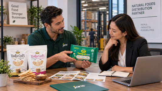 A business man and woman reviewing product brochures and packages of Royal Tempeh and Jans chips at a desk in a warehouse setting.