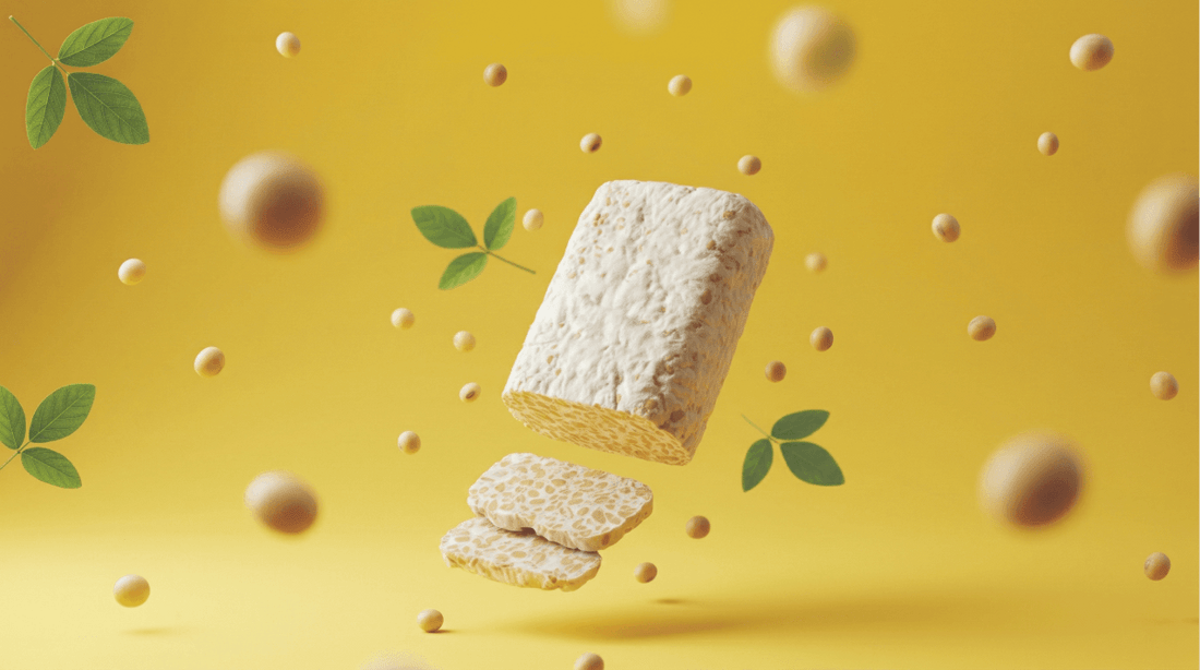A block of Royal Tempeh floating in the air with slices falling below it, surrounded by floating soybeans and green leaves on a bright yellow background.
