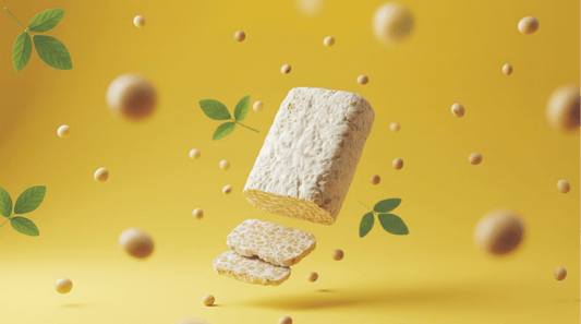 A block of Royal Tempeh floating in the air with slices falling below it, surrounded by floating soybeans and green leaves on a bright yellow background.