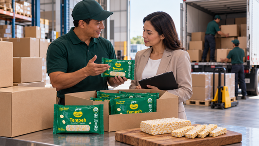 A warehouse distributor and a client reviewing packaged Royal Tempeh products and fresh slices on a wooden board next to shipping boxes.