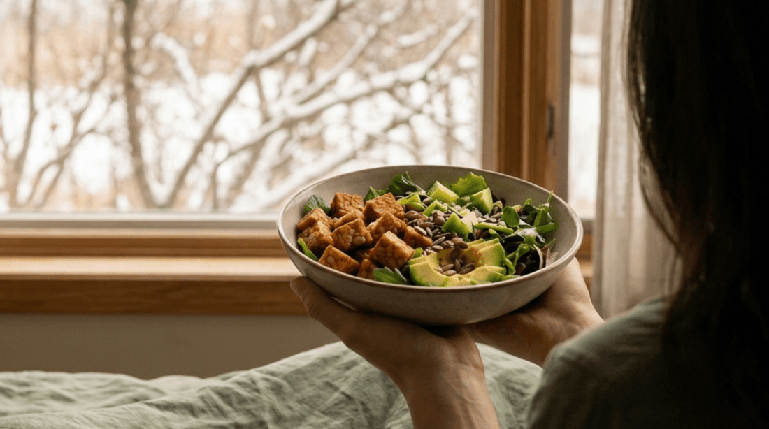A person holding a healthy bowl of cubed Royal Tempeh, avocado, and fresh greens while looking out a snowy window.