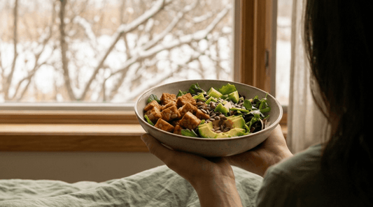 A person holding a healthy bowl of cubed Royal Tempeh, avocado, and fresh greens while looking out a snowy window.