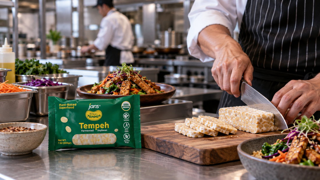A chef slicing fresh Royal Tempeh on a prep station in a professional commercial kitchen next to a retail pack and plated Asian bowls.