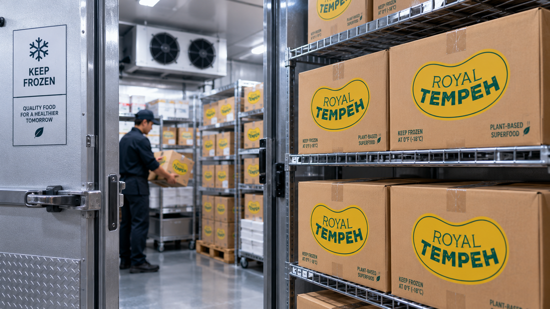 A commercial walk-in freezer stacked with boxes of Royal Tempeh on metal shelving units, with a worker organizing inventory in the background.