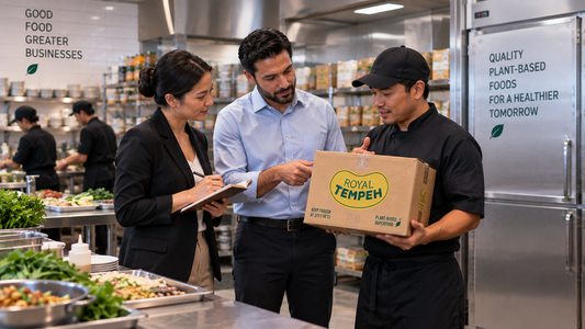 A supplier delivery representative presenting a box of Royal Tempeh to a chef and a business buyer inside a commercial kitchen preparation area.