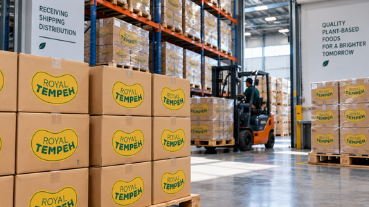 Stacked cardboard shipping boxes branded with the Royal Tempeh logo inside a modern commercial warehouse with a forklift operating in the background.