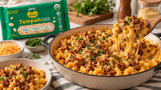 A large skillet filled with creamy vegan macaroni and cheese topped with a crispy seasoned tempeh crumble, served alongside a bowl and a green package of Royal Tempeh.